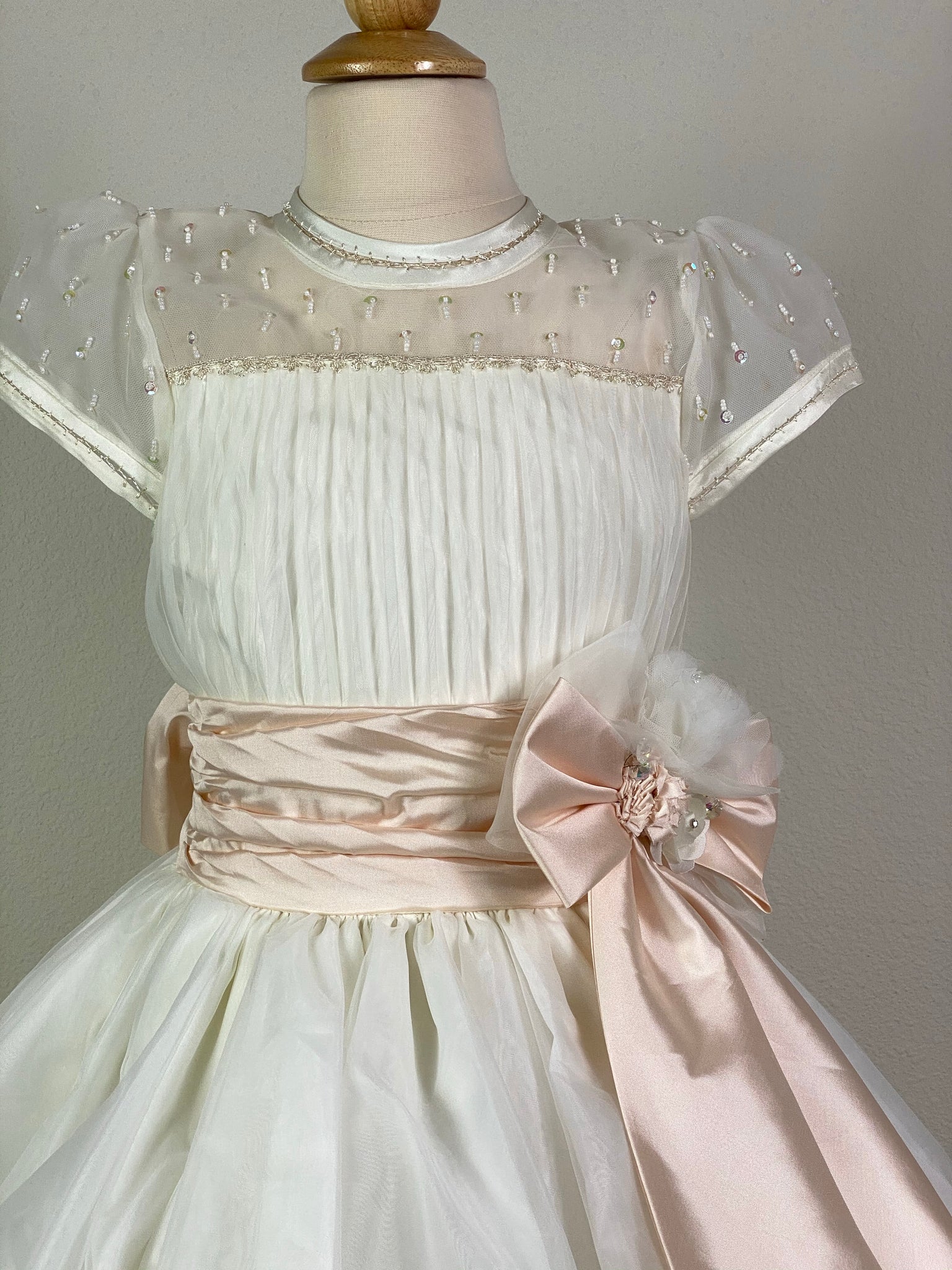 Ivory, size 10 Hand-stitched edging along sleeves and neckline Mesh and satin illusion bodice with beaded detailing and pleats Peach ruched cummerbund Large satin bow on front with floral detailing in center Meshed skirt with braided stripes along bottom Satin covered button closure Peach ribboning for large bow in back