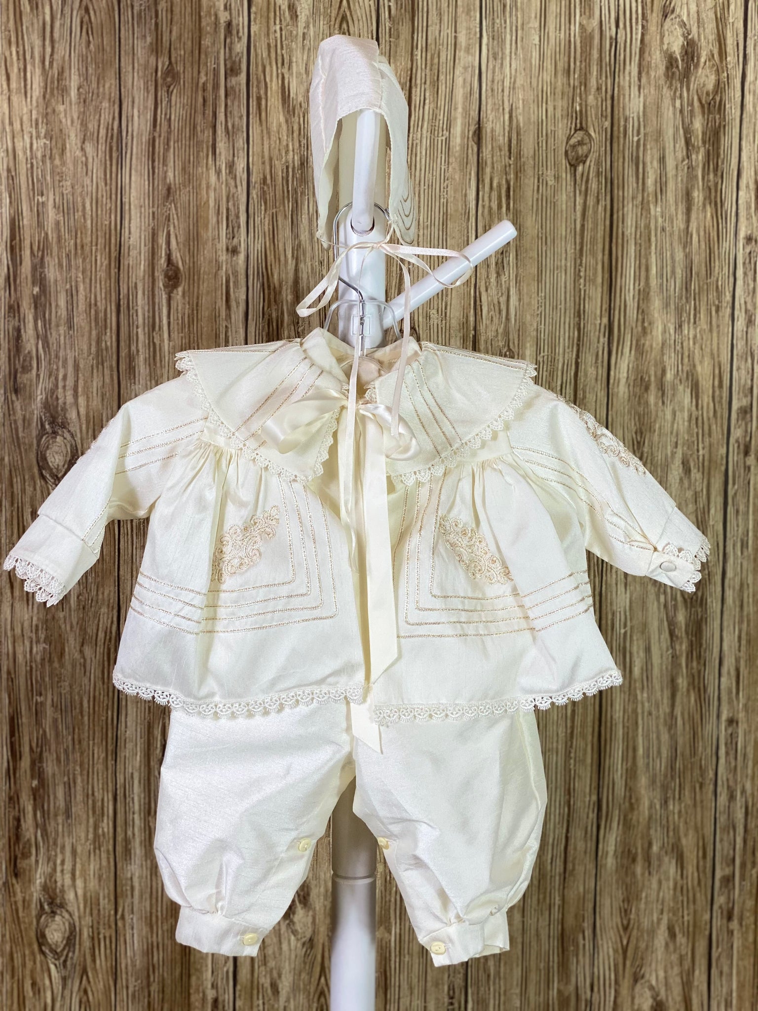 IVORY  3-piece ivory set with romper, jacket, and bonnet Solid ivory satin romper with buttons along inside of legs Collard, short sleeve romper Champagne rope detailing in L shape on jacket front Champagne rope detailing vertical along jacket collar and sleeves Embroidered appliques on jacket front Embroidered trim along jacket, collar, and sleeve edging Ribbon closure on jacket Button closure on back of romper Thin ribbon for tie on bonnet