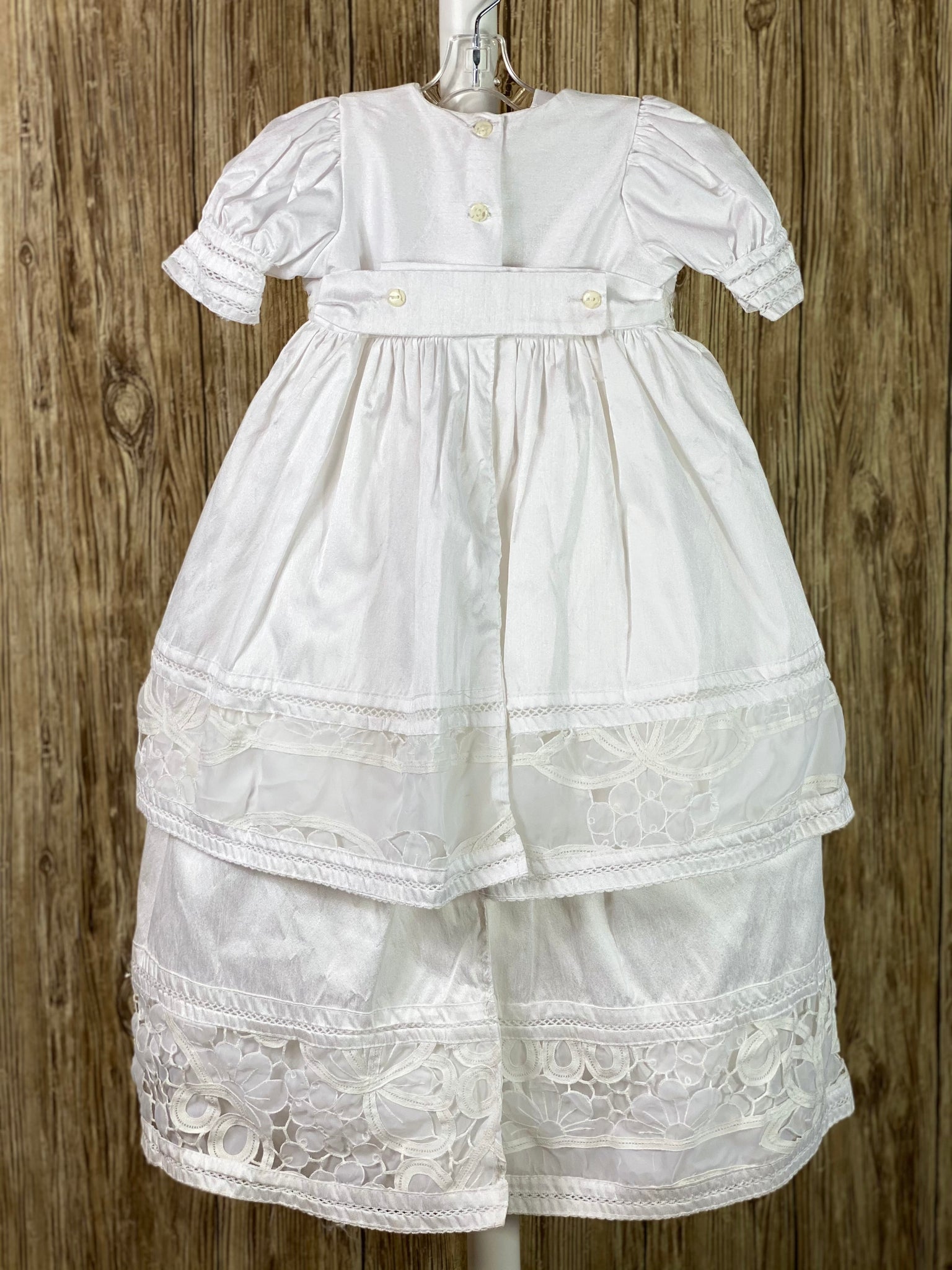 This a beautiful, one-of-a-kind baptism gown.  A lovely gown for a precious child.  White, size 24M Two layered dress Satin bodice with lace detailing Rhinestones along bodice center Flower bundle along bodice edge Puffed satin sleeves with lace trim Satin skirting with thick lace detailing Satin second layer with thick lace detailing Button closure in the back