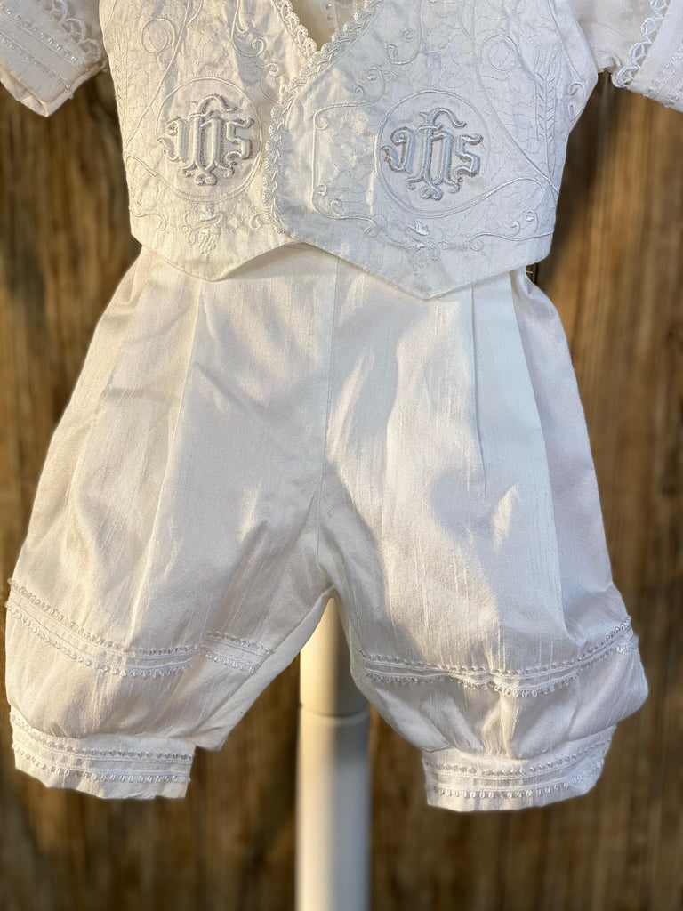 White, size 12M 4-piece set including beret, vest, shirt, suspender pants JHS symbol embroidered into vest (JHS stands for Jesus Holmium Salvatore, Latin for Jesus Savior of Mankind) Thin tasseled trim along cuffs, beret, and around pant legs Crochet trim around collar and sleeves Intricate embroidery on vest around JHS symbol Collared shirt with short sleeves Buttoning on pant cuffs Elastic banding behind pants Button closure on back of shirt