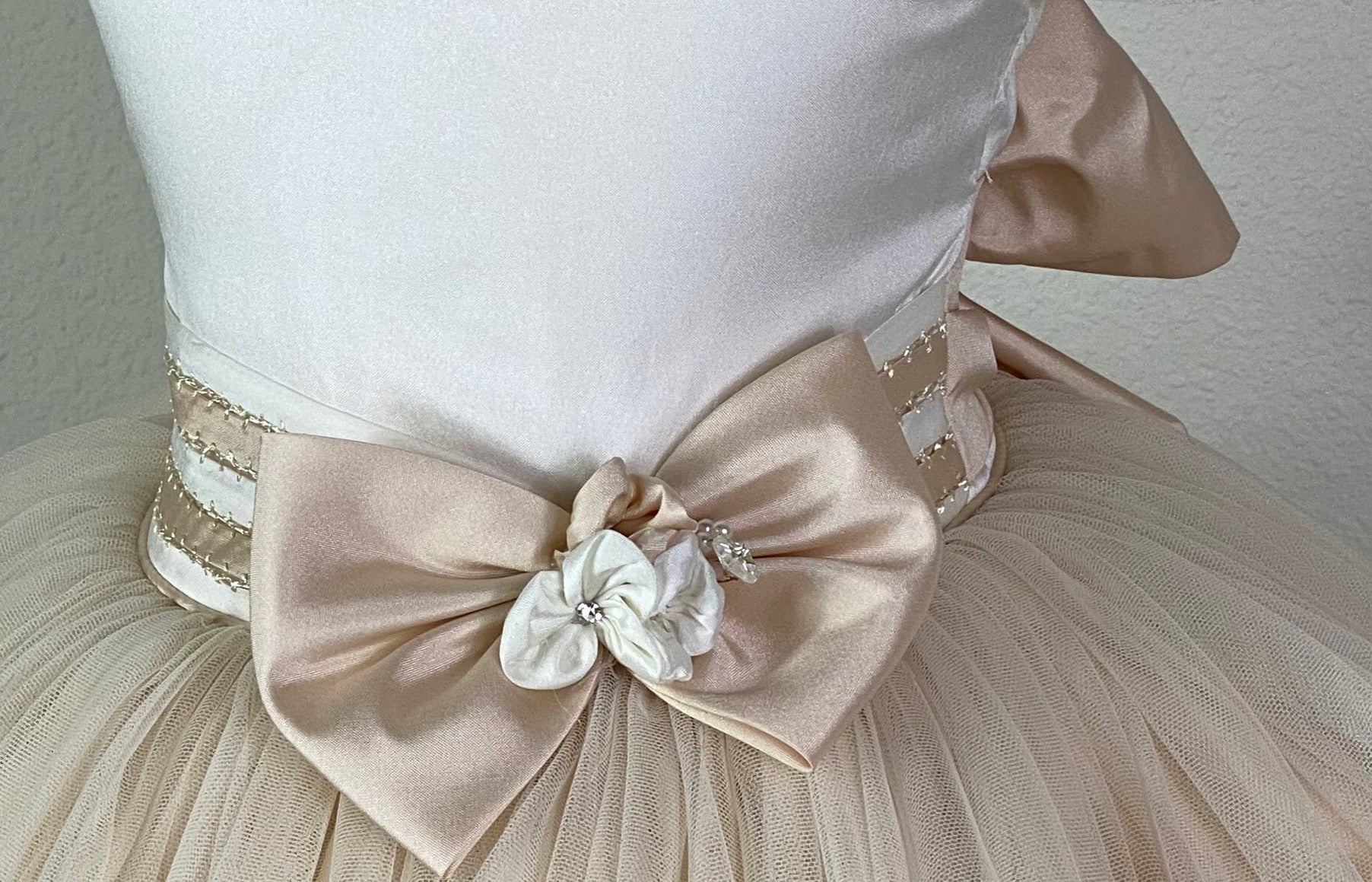 Ivory, size 2 Ivory bodice with hand stitched trim along neckline and sleeves Ruffled detailing along sleeves with hand stitched detailing Ivory and beige cummerbund with beige bow Ivory flowers in center of beige bow Tulle skirting with beige trim along edging Satin button closure Beige ribbon for bow in back Dress pictured with a petticoat Petticoat not included  Choose from a tulle, cloth, or wire for best look