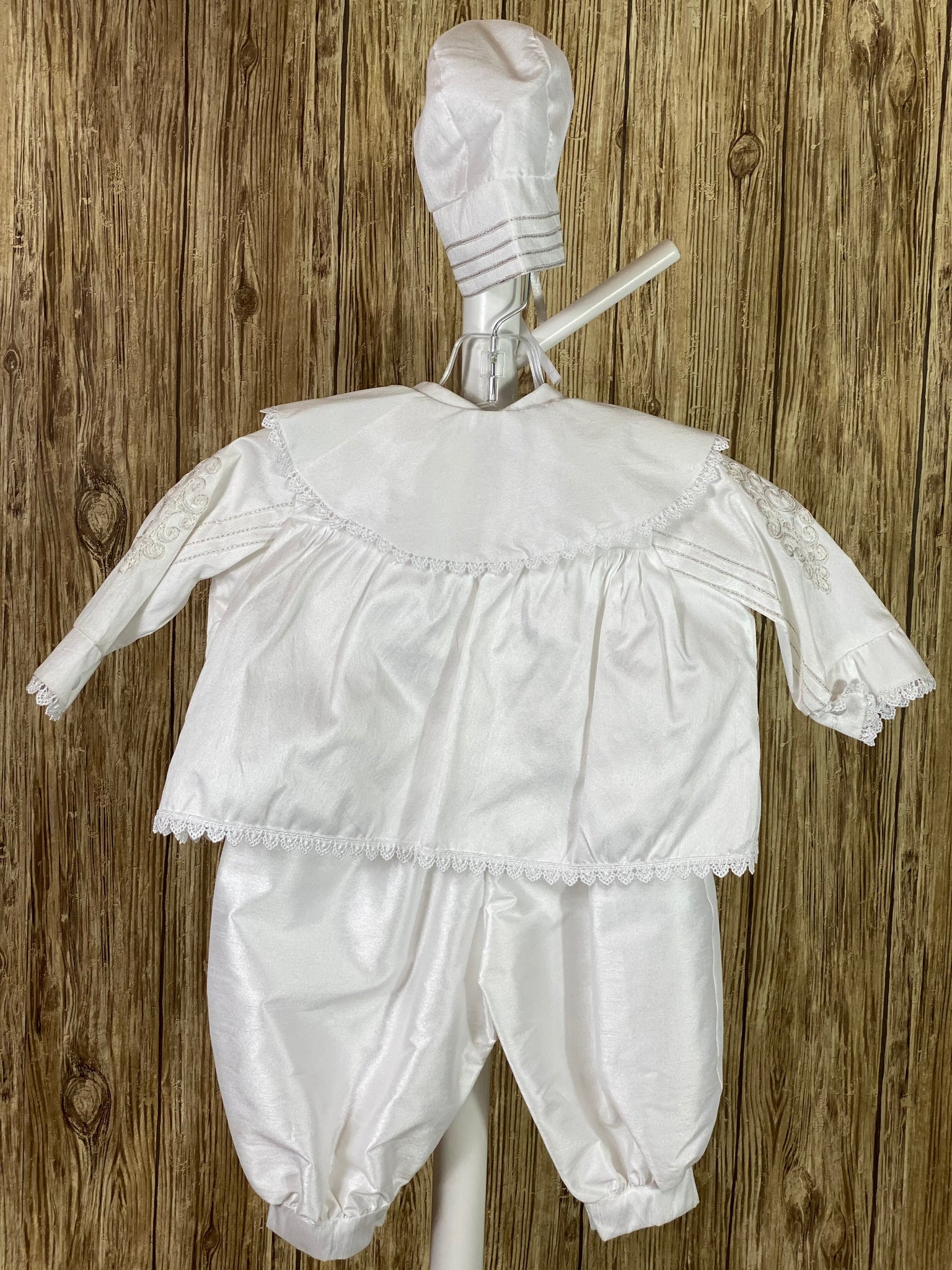 WHITE  3-piece white set with romper, jacket, and bonnet Solid white satin romper with buttons along inside of legs Collard, short sleeve romper Silver rope detailing in L shape on jacket front Silver rope detailing vertical along jacket collar and sleeves Embroidered appliques on jacket front Embroidered trim along jacket, collar, and sleeve edging Ribbon closure on jacket Button closure on back of romper Thin ribbon for tie on bonnet