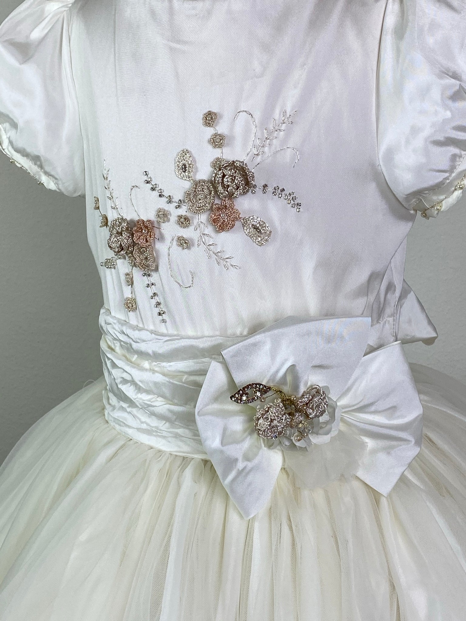 Ivory, size 10 Puffed short sleeve Hand stitched satin and pearl trim along sleeve and neckline Neutral colored embroidered flower applique along ivory satin bodice Large bow on ivory cummerbund with rhinestone and floral detailing Tulle skirt with embroidered appliques scattered Ivory satin covered button closure Ivory ribbon for large bow