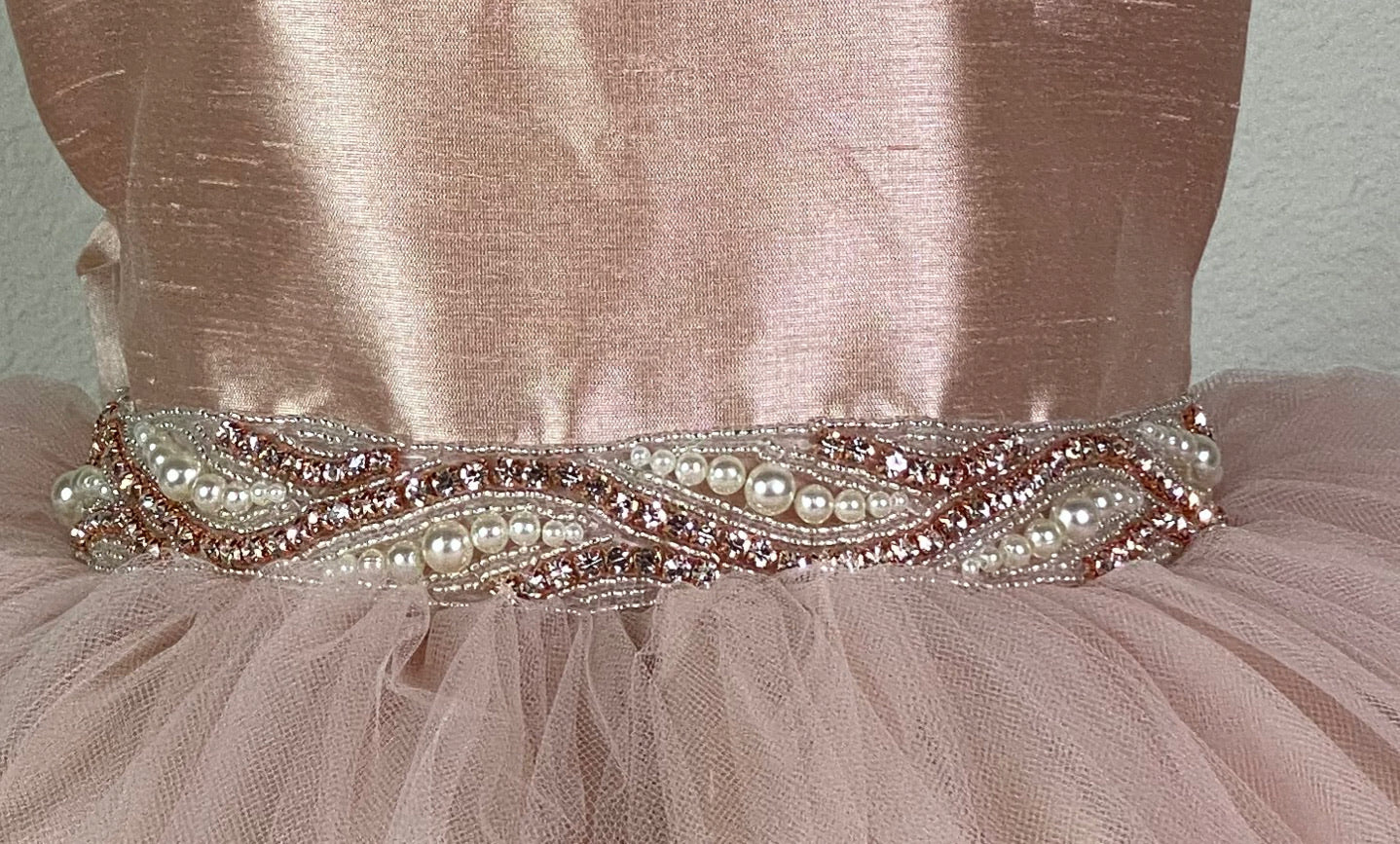 Rose gold ribbed satin bodice Rose gold pearl and rhinestone embellished band along lower bodice Satin skirting with rose gold tulle overlay Pearl button closure Satin rose gold ribbon for bow in back Dress pictured with a petticoat Petticoat not included  Choose from a tulle, cloth, or wire for best look