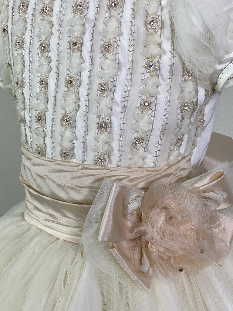 Puffed tulle short sleeve with handstitched trim along sleeve and neckline Intricate satin columns handstitched together with rhinestones Beige ruched cummerbund with floral layered large bow Leaf detailing scattered along tulle skirt Satin button closure Beige ribboning for large bow