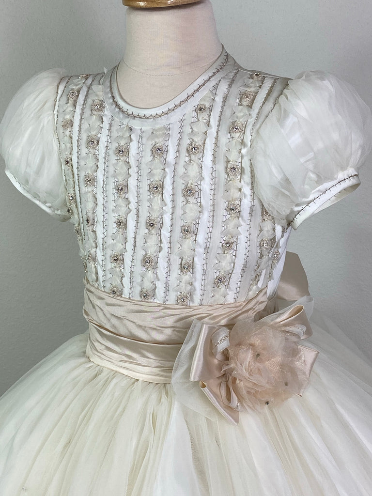 Puffed tulle short sleeve with handstitched trim along sleeve and neckline Intricate satin columns handstitched together with rhinestones Beige ruched cummerbund with floral layered large bow Leaf detailing scattered along tulle skirt Satin button closure Beige ribboning for large bow