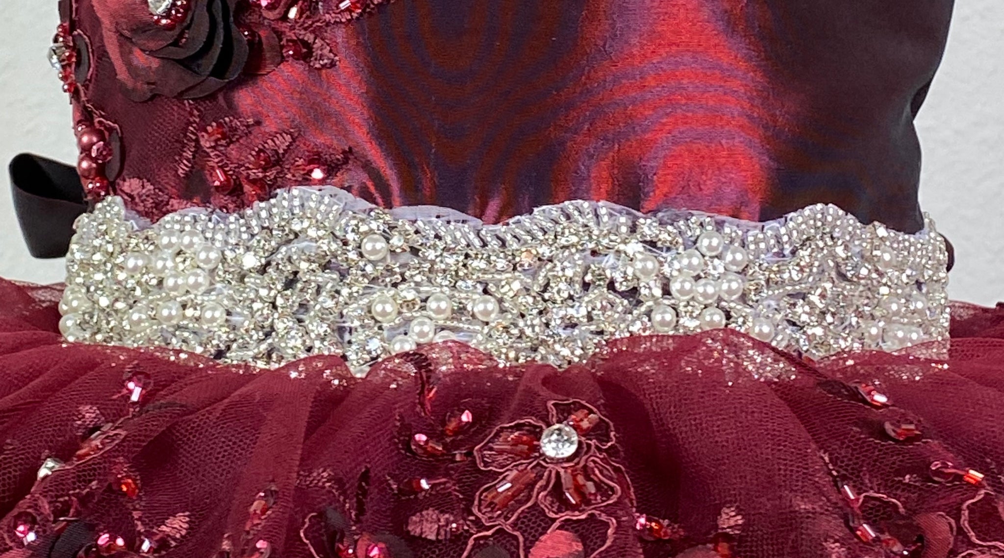 Burgundy, ivory, pink, or champagne scoop neck bodice Large flower placed on right shoulder with rhinestone detailing Embroidered flower applique under large flower Pearl and rhinestone band along lower bodice Satin skirting with tulle overlay Embroidered flower appliques along top of skirt, as well as trimming the edge of skirt Corset backing Burgundy, ivory, pink, or champagne ribbon for large bow Dress pictured with a petticoat