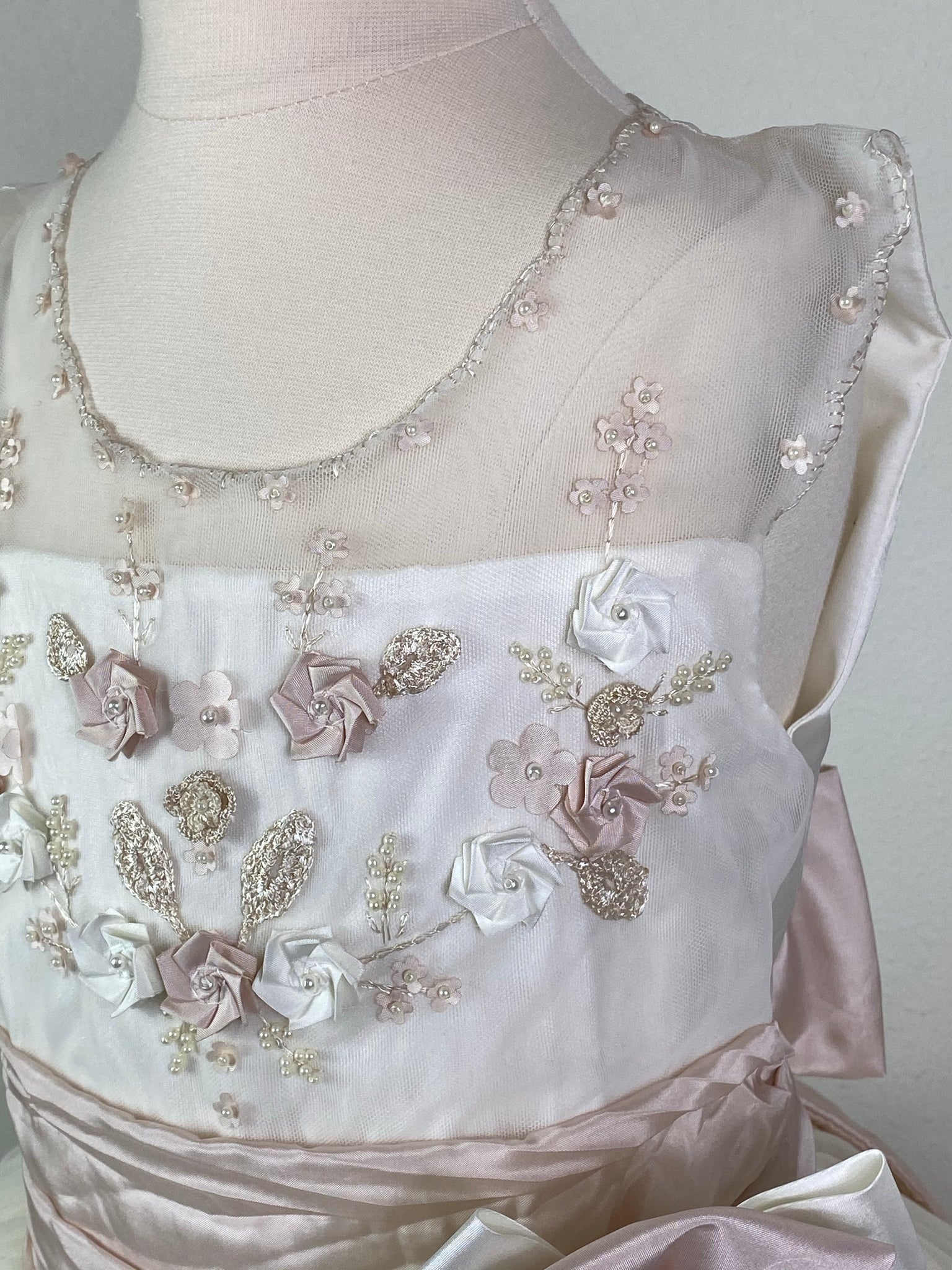 Ivory, size 10 Hand-stitched edging along arm holes and neckline Antique Rose and Ivory floral detailing with pearl center Antique Rose ruched cummerbund with pearl detailing in large bow Tulle skirt with antique rose satin trim Satin covered button closure Antique Rose ribboning for large bow Antique Rose satin trim