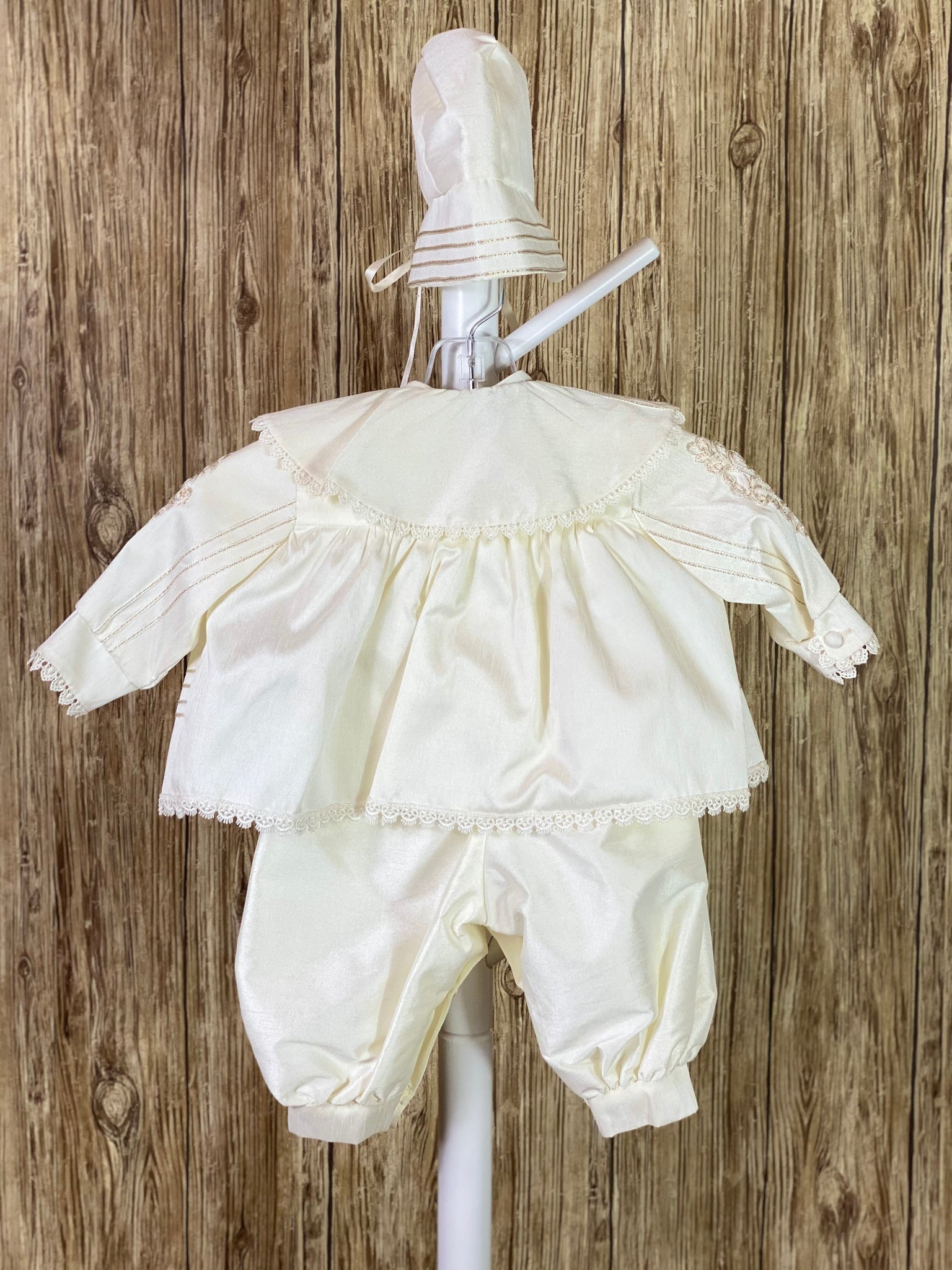 IVORY  3-piece ivory set with romper, jacket, and bonnet Solid ivory satin romper with buttons along inside of legs Collard, short sleeve romper Champagne rope detailing in L shape on jacket front Champagne rope detailing vertical along jacket collar and sleeves Embroidered appliques on jacket front Embroidered trim along jacket, collar, and sleeve edging Ribbon closure on jacket Button closure on back of romper Thin ribbon for tie on bonnet