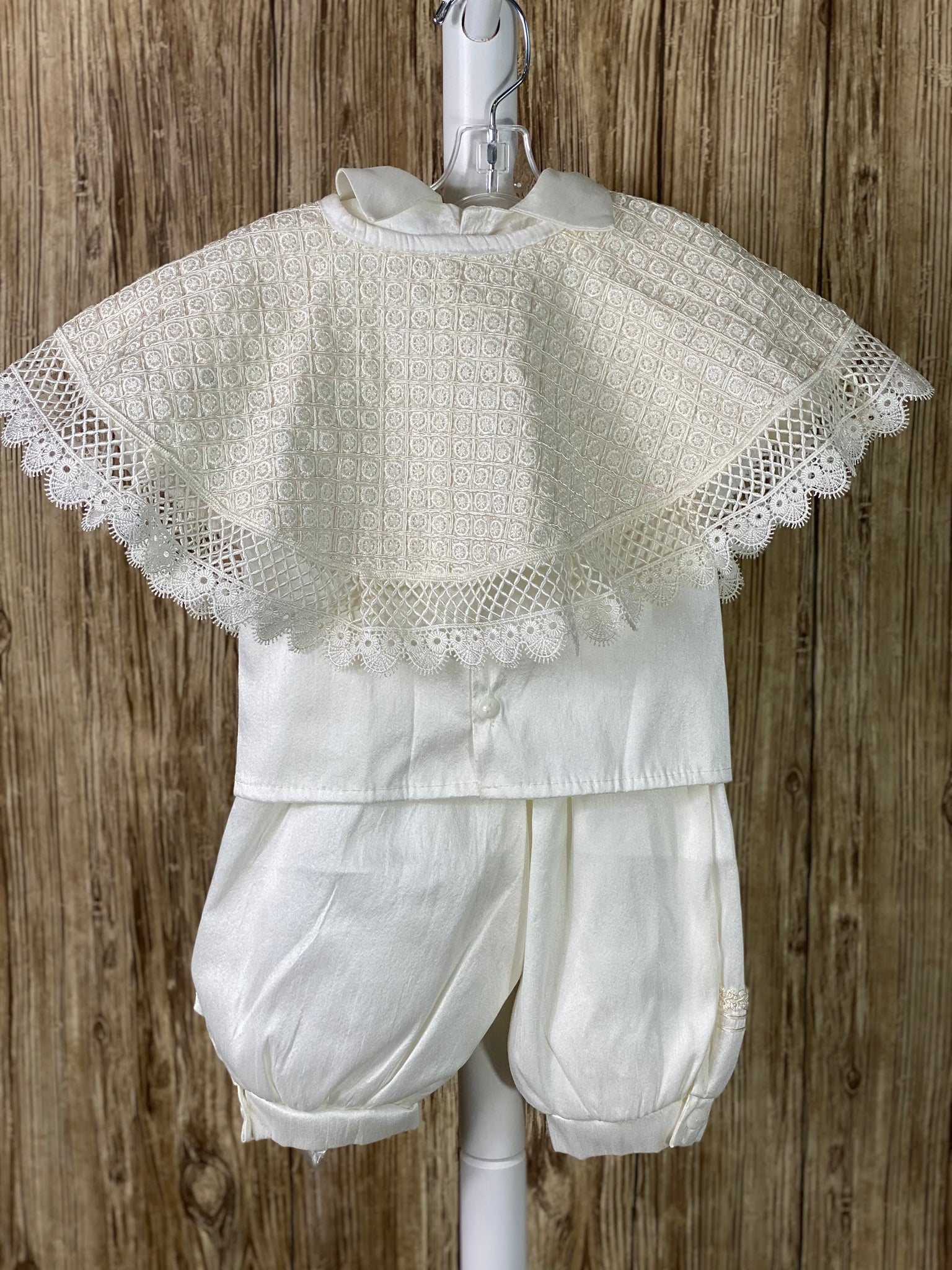 IVORY   4-piece ivory set including mozzetta, beret, shirt, suspender pants Collared romper with short sleeves Buttoning on pant cuffs Button closure on back of shirt Pin tucks overlapping diagonal with pin wheel detailing inside on mozzetta Wide embroidered lace trim around mozzetta Pearl and champagne jeweled crosses on mozzetta Rope closure on mozzetta Braided embroidered line detailing on pant, cummerbund, sleeves, beret, and down shirt center