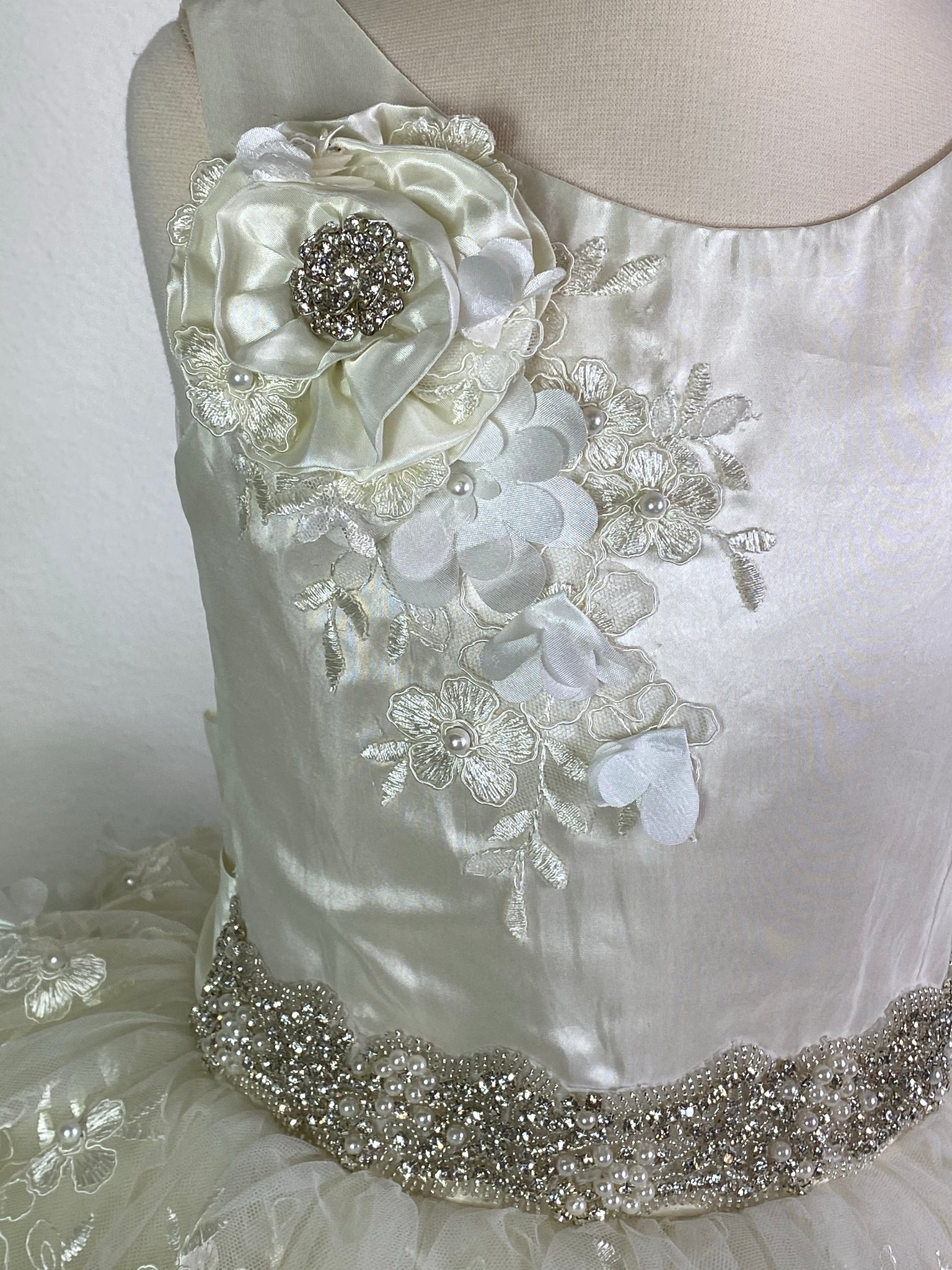 Ivory, size 8 Ivory bodice with large rhinestone flower on shoulder Pearl and rhinestone band along waist Ivory satin skirt with embroidered tulle overlay Pearl button closure Satin Ribbon for large bow