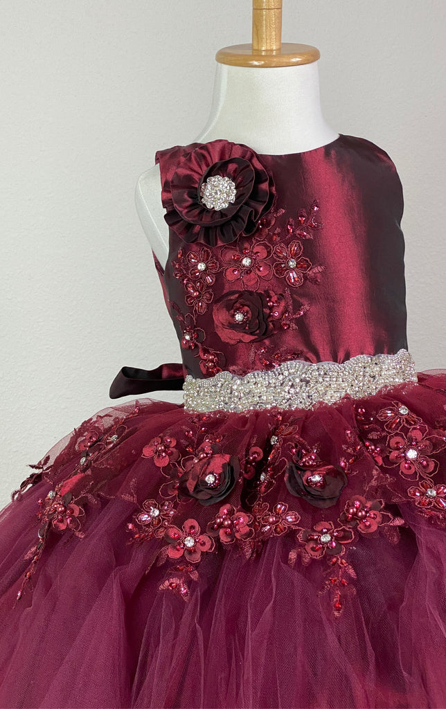 Burgundy, ivory, pink, or champagne scoop neck bodice Large flower placed on right shoulder with rhinestone detailing Embroidered flower applique under large flower Pearl and rhinestone band along lower bodice Satin skirting with tulle overlay Embroidered flower appliques along top of skirt, as well as trimming the edge of skirt Corset backing Burgundy, ivory, pink, or champagne ribbon for large bow Dress pictured with a petticoat