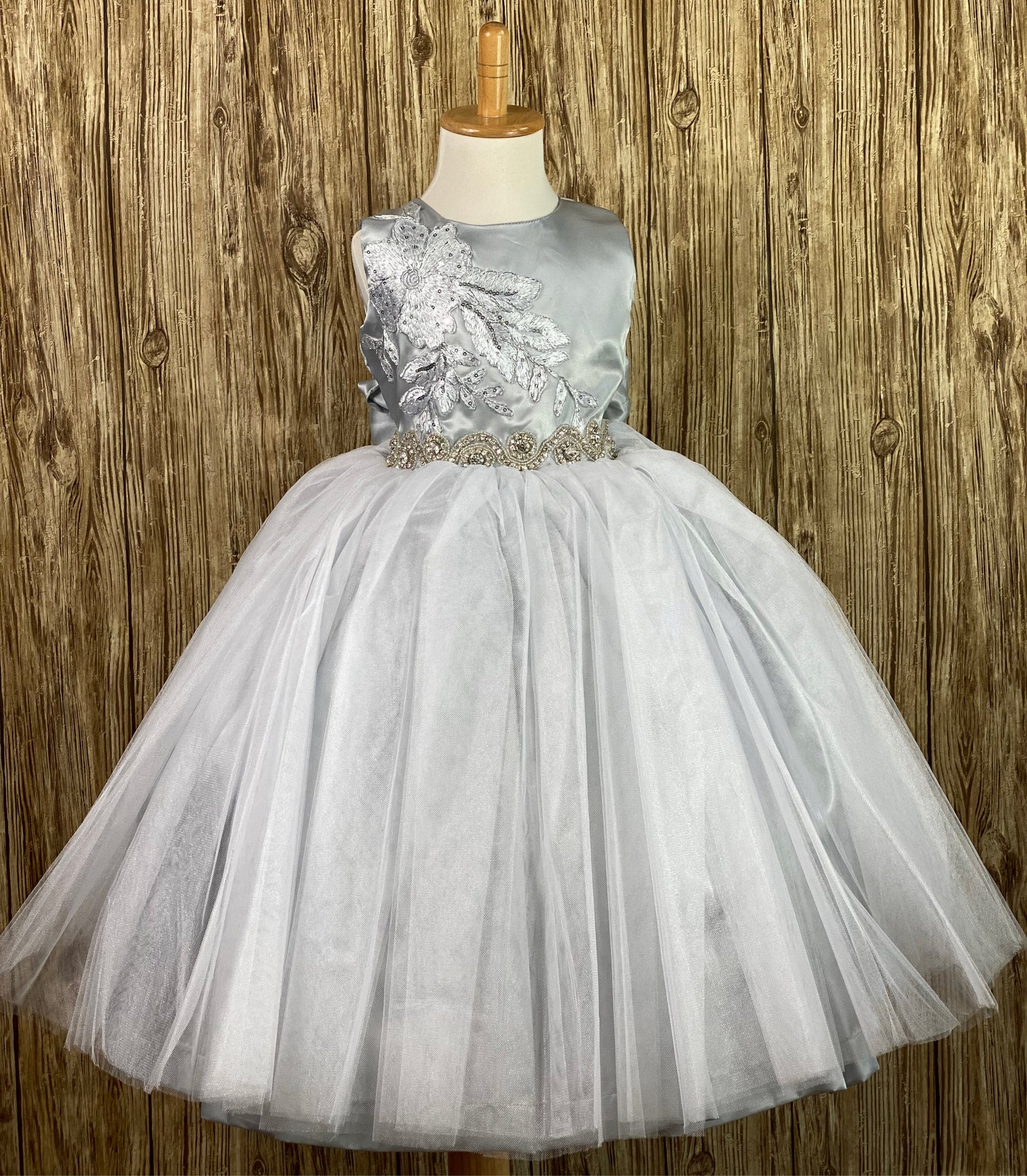 Silver, size 2 Silver bodice with large embroidered sequins and beaded flower on right side Swirled rhinestone band along lower bodice Silver satin and tulle skirting Button closure Silver ribbon for bow on back Dress pictured with a petticoat Petticoat not included  Choose from a tulle, cloth, or wire for best look
