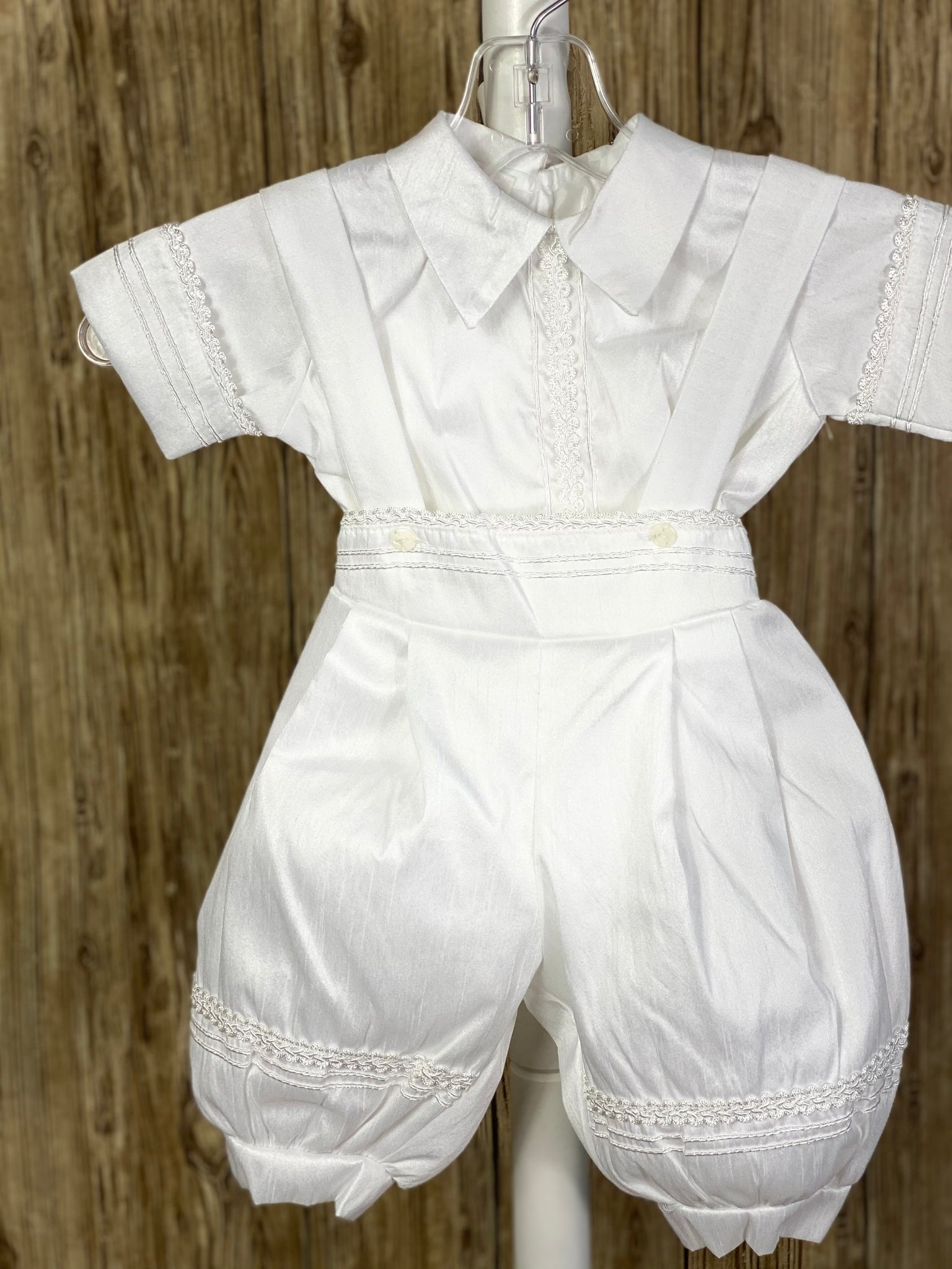 WHITE   4-piece white set including mozzetta, beret, shirt, suspender pants Collared romper with short sleeves Buttoning on pant cuffs Button closure on back of shirt Pin tucks overlapping diagonal with pin wheel detailing inside on mozzetta Wide embroidered lace trim around mozzetta Pearl and white jeweled crosses on mozzetta Rope closure on mozzetta Braided embroidered line detailing on pant, cummerbund, sleeves, beret, and down shirt center